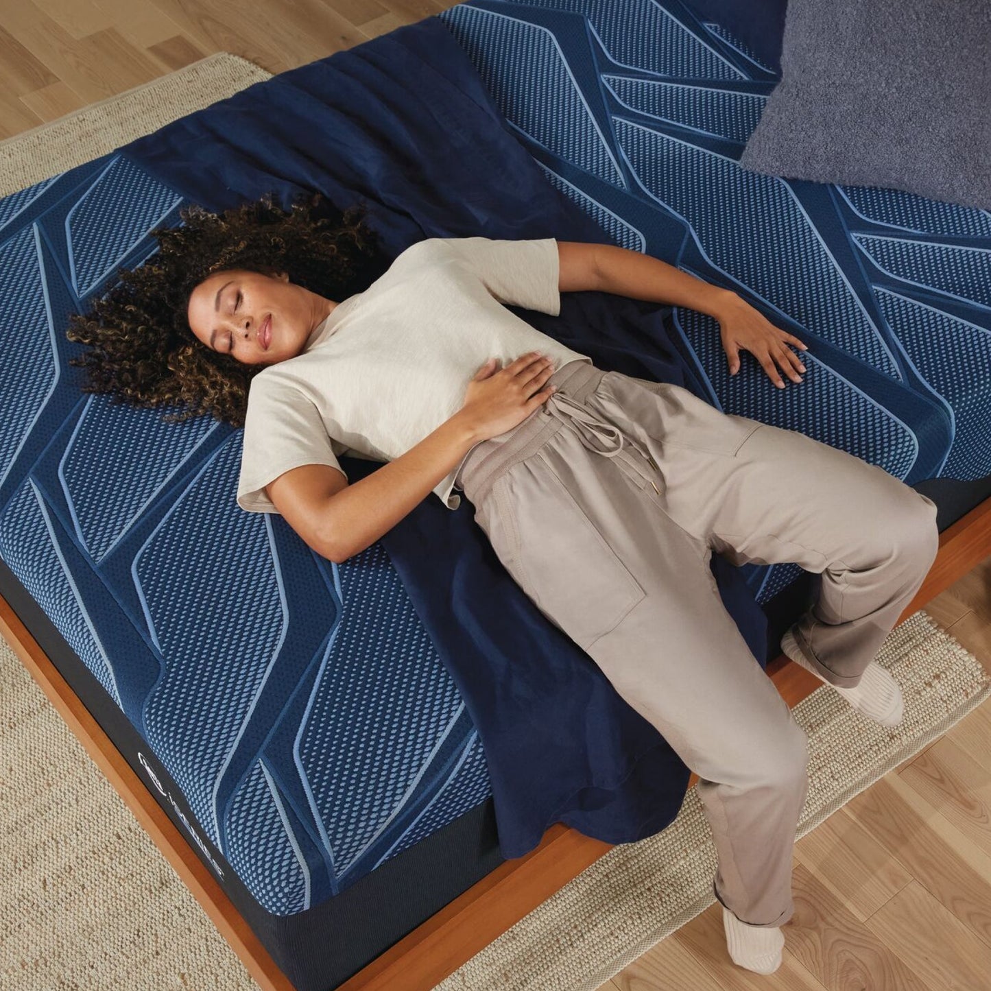 A woman laying on her back on a Serta iSeries plush blue mattress with a blue blanket under it and a blue-grey pillow at the head of the bed||feel: plush||level: Ultimate