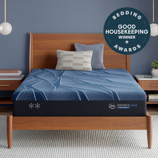 A Blue Serta® iSeries® NXG™ Mattress  on a wooden bed frame in a styled bedroom setting, featuring a blue patterned bedding cover, two pillows, a blue wall, and a 'Bedding Good Housekeeping Winner Awards' badge overlay.