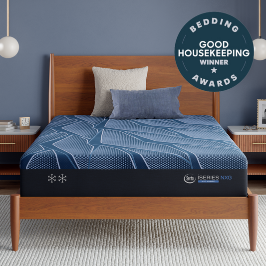 A Blue Serta® iSeries® NXG™ Mattress on a wooden bed frame in a styled bedroom setting, with a blue patterned bedding cover, two pillows, side tables, pendant lights, and a circular badge reading 'Bedding Good Housekeeping Winner Awards.' The mattress brand mark 'iseries' is visible near the bottom edge.