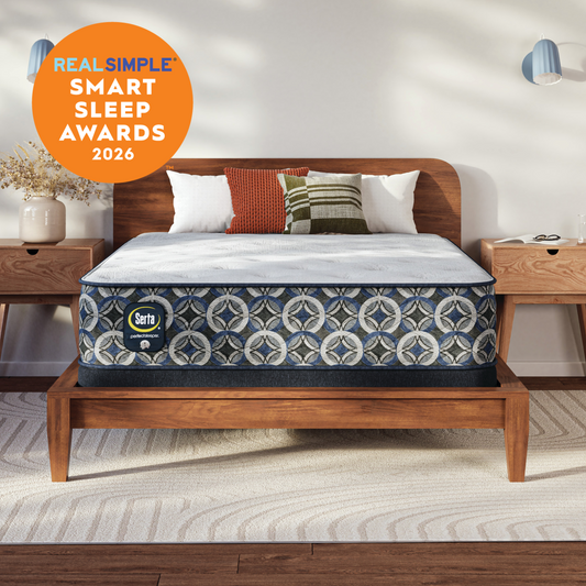 A Serta Perfect Sleeper plush mattress on a wooden bed frame in a styled bedroom setting, with decorative pillows, bedside tables, wall sconces, and a beige rug. An orange circular badge in the top left reads 'REAL SIMPLE SMART SLEEP AWARDS 2026'.