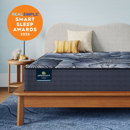 A Perfect Sleeper X Hybrid mattress on a wooden bed frame in a styled bedroom scene with a side table, pillows, rug, and a light blue wall. An orange badge in the top left reads 'REAL SIMPLE SMART SLEEP AWARDS 2026.