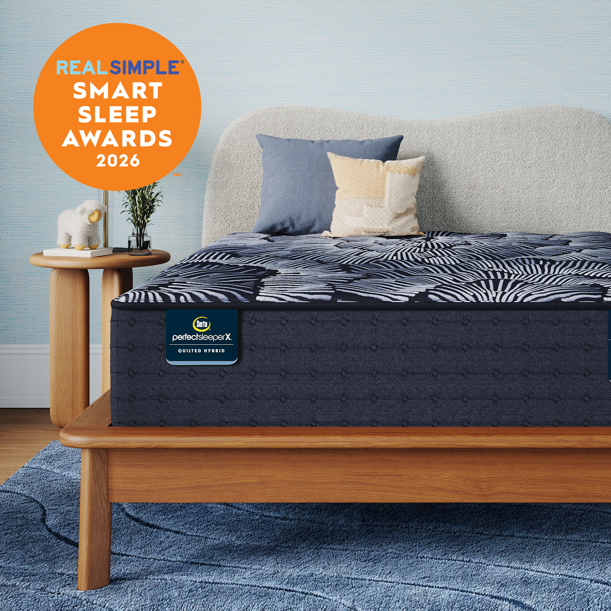 A Perfect Sleeper X Quilted Hybrid Excelled Plush mattress with a dark pattern and  matching pillow shams styled in a bedroom setting, with a light wood bed frame, blue rug, side table, and a large orange badge reading 'REAL SIMPLE SMART SLEEP AWARDS 2026'. The mattress label shows 'perfectscore X' and 'BETTER START'.