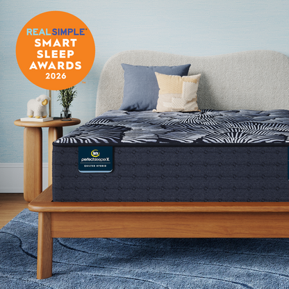 A Perfect Sleeper X Quilted Hybrid Excelled Plush mattress with a dark pattern and  matching pillow shams styled in a bedroom setting, with a light wood bed frame, blue rug, side table, and a large orange badge reading 'REAL SIMPLE SMART SLEEP AWARDS 2026'. The mattress label shows 'perfectscore X' and 'BETTER START'.