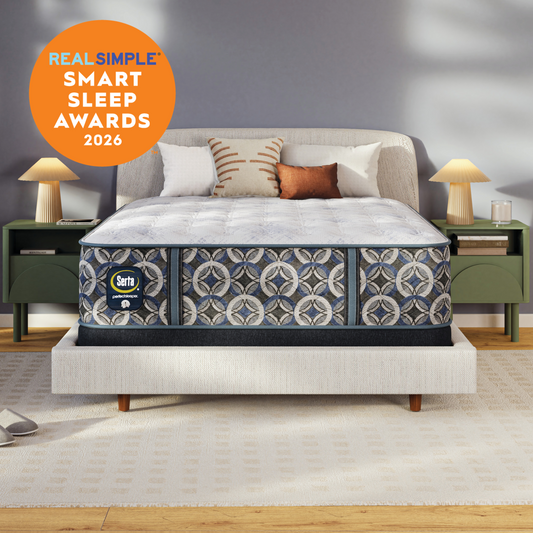 A Serta perfect sleeper plush mattress with a pattern matching upholstered base in a styled bedroom scene, with two green nightstands, lamps, pillows, and a gray wall. An orange circular badge in the upper left reads 'REAL SIMPLE SMART SLEEP AWARDS 2026.
