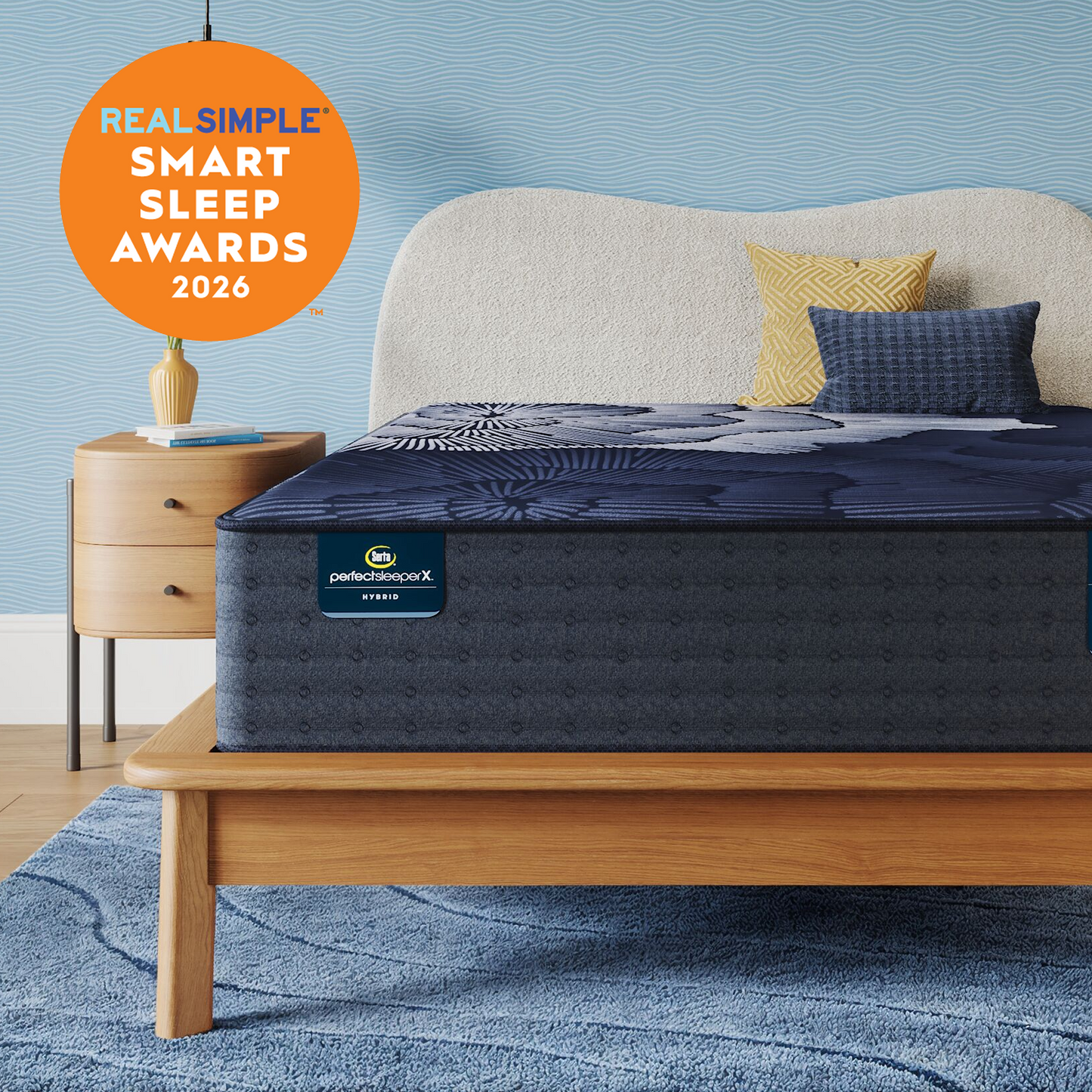 A Perfect Sleeper X Hybrid mattress with a dark navy pattern on a light wood bed frame, styled with a cream upholstered headboard, yellow and blue pillows, a wooden nightstand, and a blue textured rug. An orange circular badge in the top left reads 'REAL SIMPLE SMART SLEEP AWARDS 2026'.