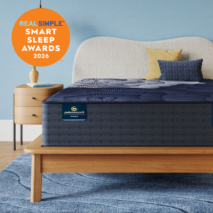 A Perfect Sleeper X Hybrid mattress with a dark navy pattern on a light wood bed frame, styled with a cream upholstered headboard, yellow and blue pillows, a wooden nightstand, and a blue textured rug. An orange circular badge in the top left reads 'REAL SIMPLE SMART SLEEP AWARDS 2026'.