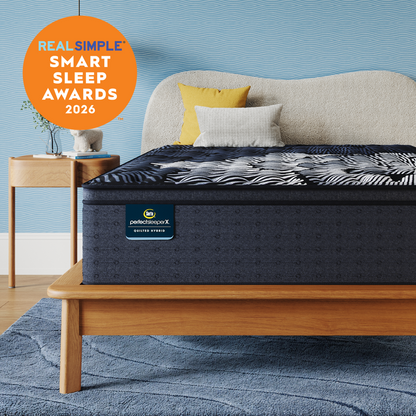 A Queen-sized Perfect Sleeper X Quilted Hybrid mattress with a dark navy and white patterned top surface is shown on a wooden bed frame in a styled bedroom. The background wall is textured light blue, with a beige upholstered headboard, two accent pillows (one yellow, one white), a matching blue rug, and a small wooden side table.