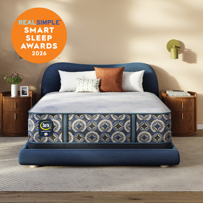 A Serta Perfect Sleeper Hybrid Firm mattress patterned shown in a styled bedroom setting with wooden nightstands, a wall lamp, and a beige wall. An orange circular badge in the top left reads 'REAL SIMPLE SMART SLEEP AWARDS 2026'