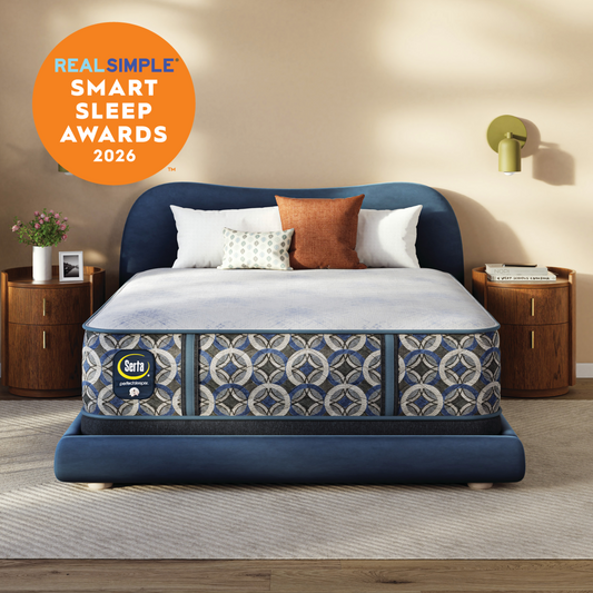A Serta Perfect Sleeper Hybrid Firm mattress patterned shown in a styled bedroom setting with wooden nightstands, a wall lamp, and a beige wall. An orange circular badge in the top left reads 'REAL SIMPLE SMART SLEEP AWARDS 2026'