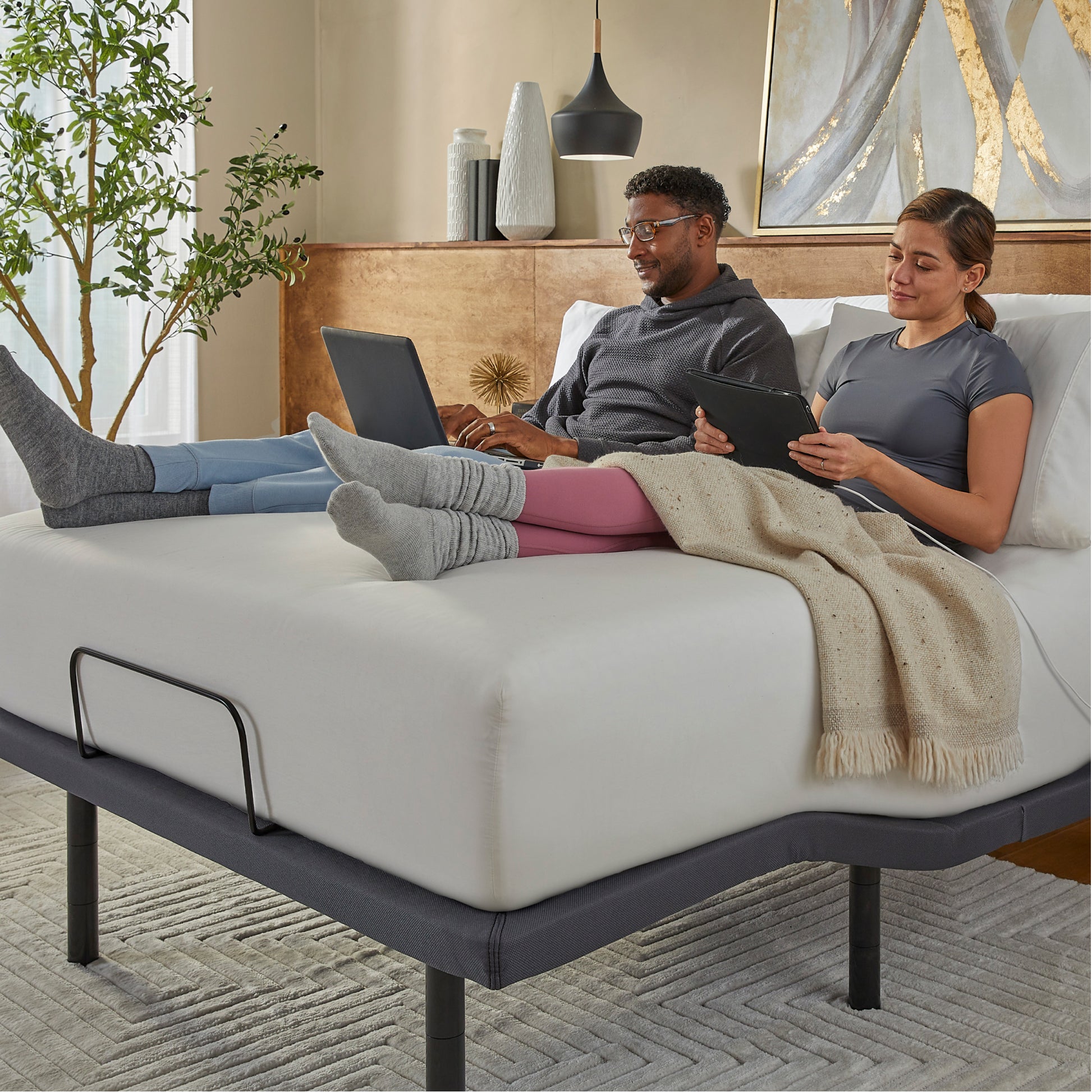 Two people using laptops on a mattress on top of a Baselogic Pearl adjustable base in a cozy room.